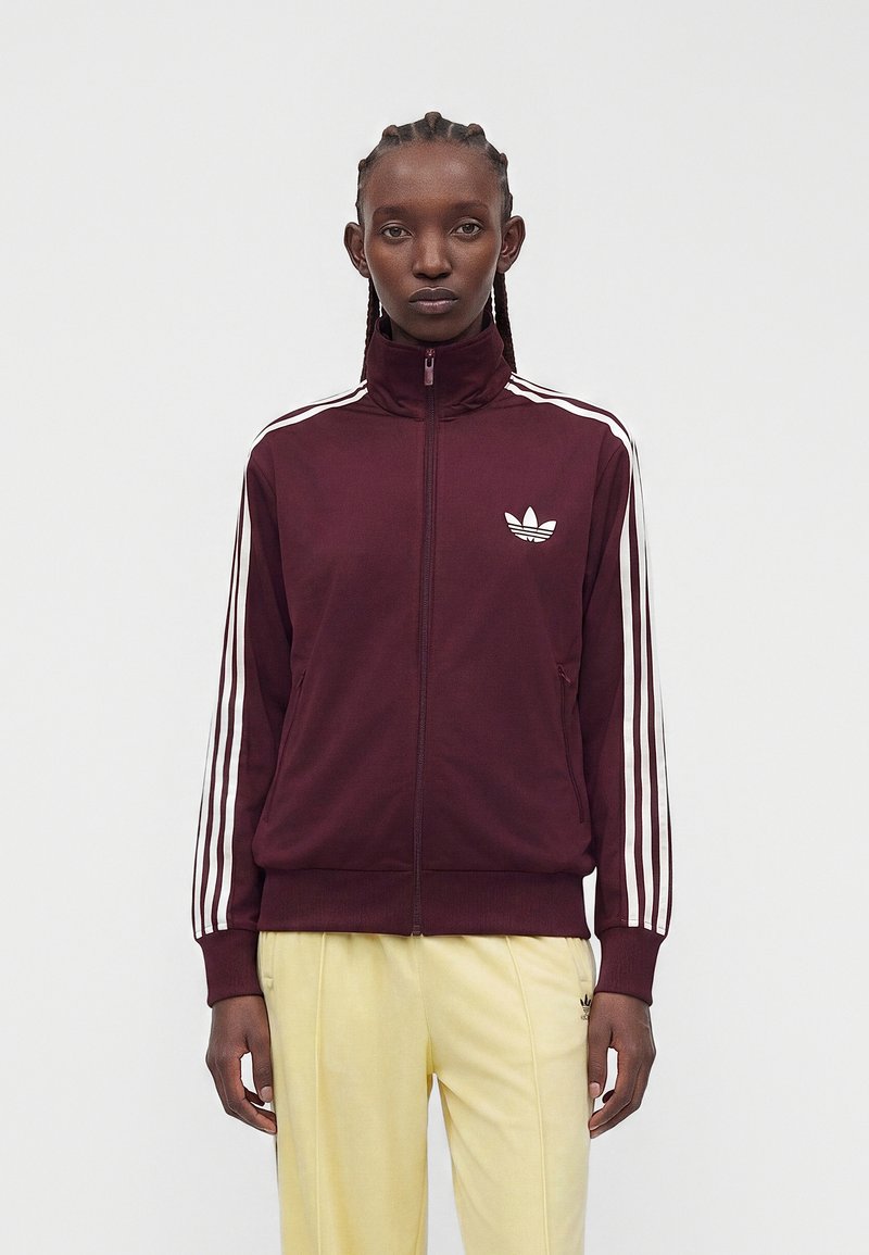 Άτομο με πλεξούδες που φορά ένα μπορντό Adidas τζάκετ με λευκές ρίγες και λογότυπα, συνδυασμένο με ανοιχτό κίτρινο παντελόνι, στέκεται μπροστά σε απλό φόντο.
