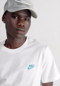 Vitt bomullströja med en turkos Nike-logotyp på vänster bröst, kombinerad med en silverfärgad keps och placerad mot en neutral bakgrund.