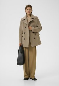 Cappotto di lana beige con ampio colletto, design doppiopetto e bottoni neri. Abbinato a pantaloni beige larghi e una borsa tote nera.