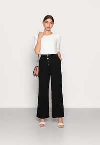 Witte getextureerde blouse met korte pofmouwen, gecombineerd met hoge zwarte wijdvallende broek met vijf knopen en zijzakken.