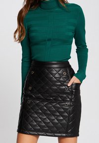 Femme portant un pull à col roulé côtelé vert et une jupe en cuir matelassé noire avec des détails de boutons dorés, la main dans la poche de la jupe.