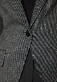 Gros plan sur un blazer gris à rayures fines boutonné sur un vêtement noir, mettant en valeur la texture du tissu et les détails du revers.