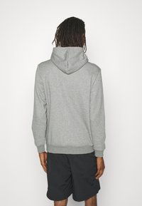 Sudadera con capucha gris de textura suave, con puños y dobladillo acanalados, y un bolsillo tipo canguro. Lleva pantalones cortos oscuros, vista desde atrás.