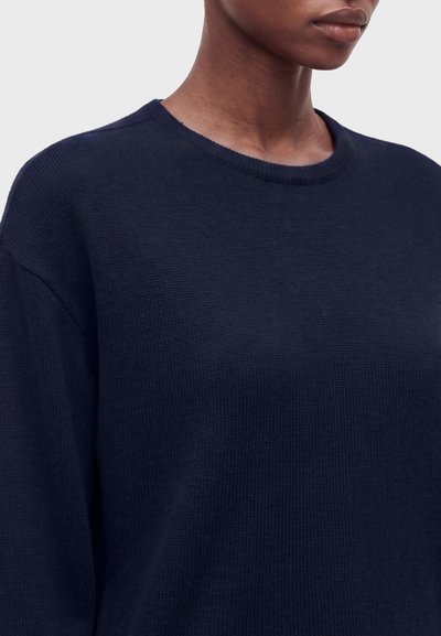Femme portant un pull en maille bleu marine foncé à col rond, montrant les épaules et une partie du cou sur un fond uni.
