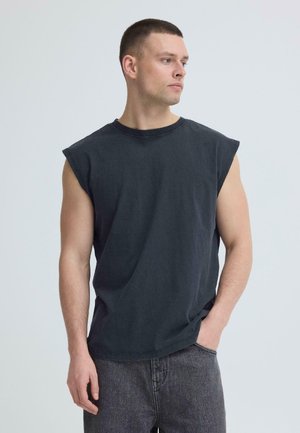 Jeune homme portant un débardeur noir et un jean gris, debout avec la main gauche dans la poche, regardant vers la droite sur un fond clair uni.