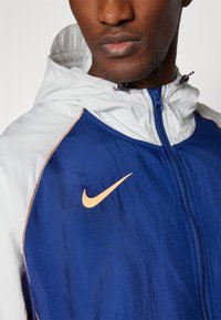Veste Nike arborant un corps bleu avec des accents blancs, une fermeture éclair et un logo orange. Le matériau semble léger et texturé.