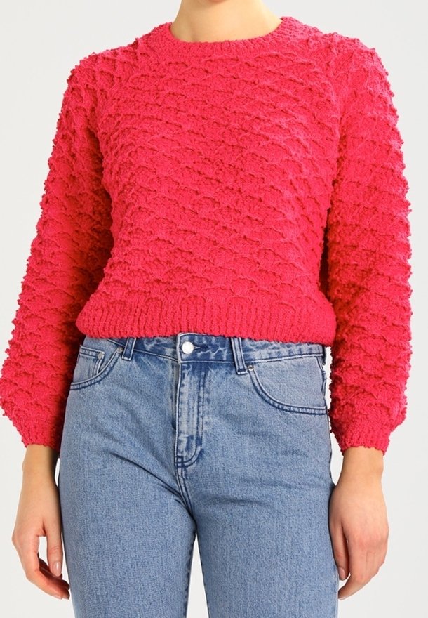 Personne portant un pull en tricot rose vif texturé et un jean bleu clair taille haute, debout avec les bras détendus.