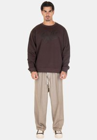 Felpa marrone con un logo sottile, abbinata a pantaloni beige a pieghe e sneakers in bianco e nero. Il modello è in piedi su uno sfondo neutro.