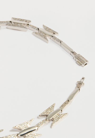 Ogrlica v srebrni barvi z ostrimi povezavami, izmenično sestavljenimi iz gladkega kovinskega dela in bleščečih kristalov, oblikovana v vzorcu medsebojnega prepletanja.