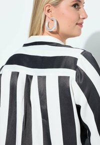 Černobílá pruhovaná blůza se saténovým povrchem, s límečkem a plisovanými detaily na zádech. Viditelné velké kruhové stříbrné náušnice.