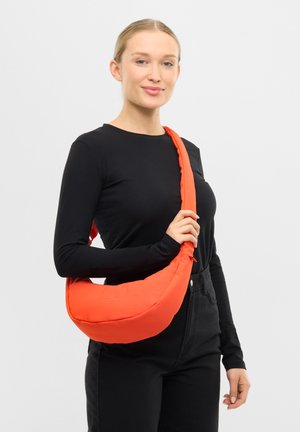 Frau trägt schwarzen langärmligen Oberteil und Hose, hält eine leuchtend orangefarbene, halbmondförmige Schultertasche vor schlichtem hellem Hintergrund.