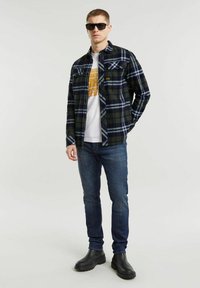 Gevlochten flanellen overhemd in marineblauw en groen, gedragen over een wit T-shirt met opdruk, gecombineerd met donkerblauwe jeans en zwarte enkellaarzen, met een zonnebril.