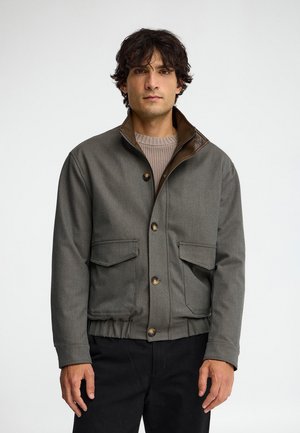 Uomo con capelli scuri e ricci indossa una giacca grigia con bottoni e tasche frontali sopra un maglione beige e pantaloni neri, in piedi su sfondo bianco.