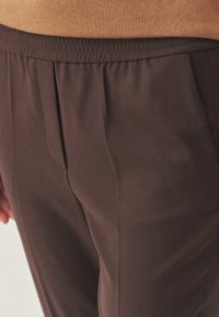 Nahaufnahme von dunkelbraunen elastischen Hosen mit einem hellbraunen Oberteil, das die Vorderfalten und die Detailansicht der Seitentaschen zeigt.