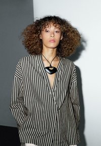 Striped blazer with a relaxed fit, featuring black and white vertical stripes, paired with a bold black pendant necklace on a thin cord.