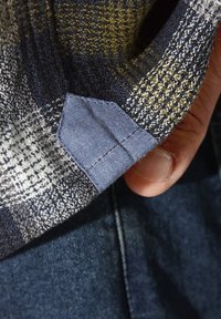 Gewebter Hemdenstoff mit kariertem Muster in Dunkelblau, Senf und Weiß. Ein blauer Textilflicken ist nahe dem Saum sichtbar.