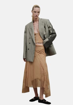 Kobieta z uczesanymi do tyłu włosami, ubrana w oversize'ową szarą marynarkę, beżowy gorset bez ramiączek, przezroczystą asymetryczną beżową spódnicę oraz czarne płaskie buty.