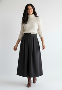 Kremowy prążkowany golf w połączeniu z czarną plisowaną spódnicą z wysokim stanem i okrągłym paskiem jako akcentem. Brązowe botki do kostek dopełniają stylizację.