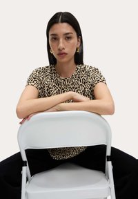 T-shirt con stampa leopardata in beige e nero, con maniche corte e scollatura a girocollo. Il modello è seduto su una sedia pieghevole bianca.