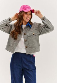 Šedá skrátená bunda s gombíkovými akcentmi, dvoma prednými vreckami, modrým golierom, skombinovaná s námorníckymi nohavicami a jasne ružovou čiapkou.