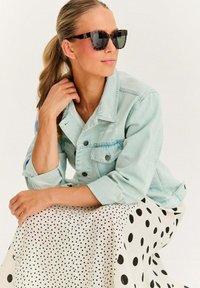 Kobieta z blond końskim ogonem, nosząca okulary w tortoise shell, jasną dżinsową kurtkę i białą spódnicę w czarne kropki, siedząca z ręką przy szyi.