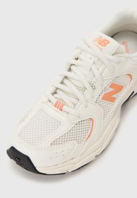 Hvite mesh løpesko med oransje detaljer, avrundet tå og polstret såle. Har snøring og logo på tungen og siden.