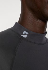 Fekete atlétikai ing magas nyakkal, sima anyagból készült. A galléron egy szürke logó található, valamint kontrasztos varrások.