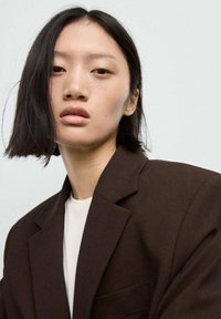 Blazer marrone con revers netti, indossato sopra una maglietta bianca. I capelli sono scuri e lisci, inquadrano un'espressione neutra. Sfondo minimalista.