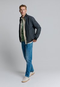 Grijs bomberjack met een ribbelkraag, gelaagd over een groene blouse en een beige onderhemd. G gedragen met blauwe spijkerbroeken en beige sneakers.