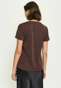 T-shirt corto marrone a maniche corte con scollatura a girocollo. Tessuto morbido e liscio, vestibilità rilassata, mostrato da dietro, abbinato a una gonna nera.