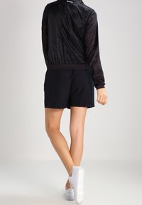 Czarna lekka kurtka bomberka z prążkowanymi mankietami, zestawiona z czarnymi szortami i białymi sneakersami, prezentująca gładką fakturę i prosty design.