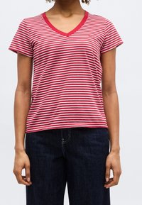 Femme portant un t-shirt rouge et blanc à rayures horizontales, col en V et manches courtes, et un jean bleu foncé, debout devant un fond uni.