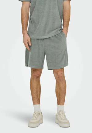 Mężczyzna noszący szare szorty z frotty i pasującą koszulkę, białe skarpetki oraz beżowe tenisówki, stoi z jedną ręką w kieszeni.