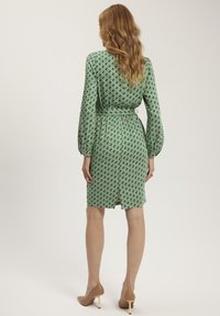 Zielona sukienka w geometryczny wzór z długimi rękawami, talią wciętą i rozcięciem z tyłu. Materiał ma wzór w diamenty. Beżowe, wysokie obcasy.