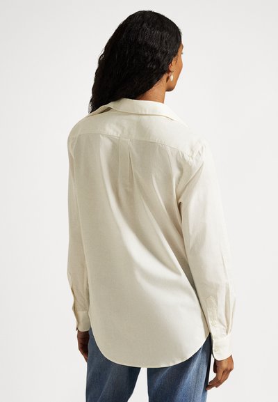 Kvinne med langt krøllete hår iført en kremfarget langermet skjorte og blå jeans, stående med ryggen mot kameraet foran en ensfarget hvit bakgrunn.