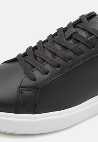 Baskets en cuir noir à finition lisse, dotées de lacets plats noirs, d'une languette rembourrée et d'une semelle en caoutchouc blanche. Détail de coutures latérales visible.