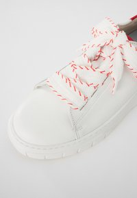 Baskets blanches en cuir avec un bout arrondi, dotées de lacets blancs avec des surpiqûres rouges et d'une semelle en caoutchouc texturé.