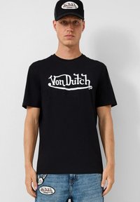 Juodas medvilninis trumpas rankovės marškinėliai su baltu "Von Dutch" logotipu. Sušalti su šviesiai mėlynomis džinsomis, ant kišenių esant logotipo pleistrams.