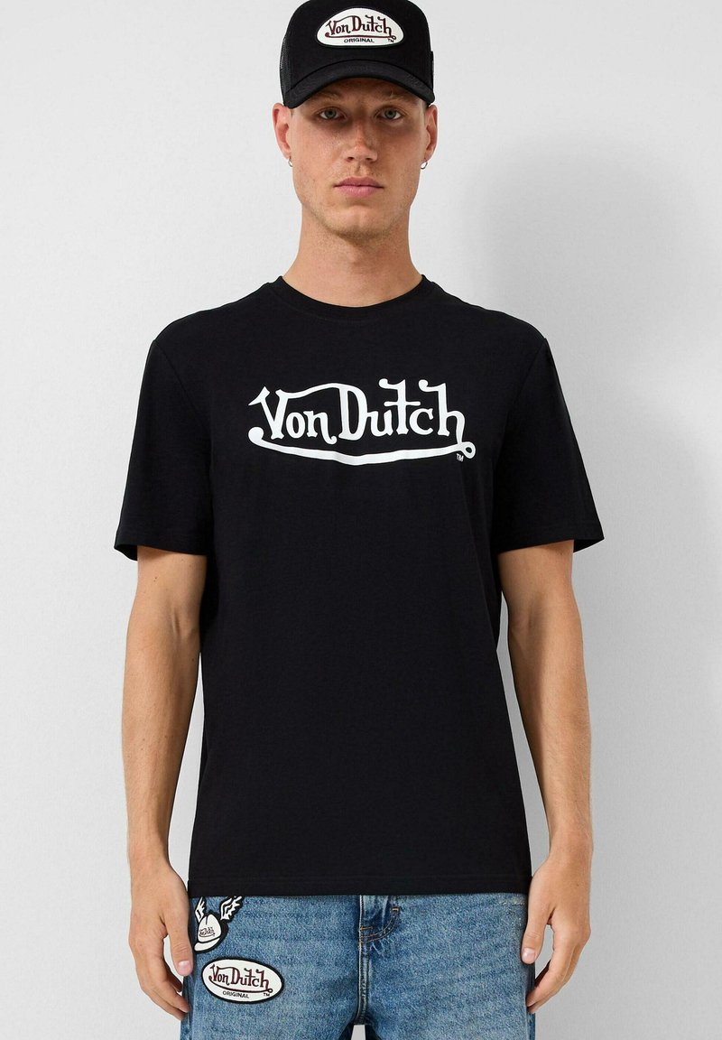 Juodas medvilninis trumpas rankovės marškinėliai su baltu "Von Dutch" logotipu. Sušalti su šviesiai mėlynomis džinsomis, ant kišenių esant logotipo pleistrams.