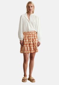 Weißes, langärmliges Button-Down-Hemd und Rock mit orangefarbenem Muster und floralem Design; getragen mit beigen, strukturierten Slipper-Sandalen.