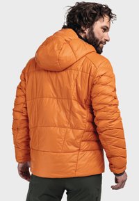 Orangene Pufferjacke mit Kapuze, die über ein gestepptes Design, eine glänzende Oberfläche und verstellbare Bündchen verfügt. Von hinten betrachtet, wird sie mit dunklen Hosen kombiniert.