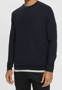 Maglione a maniche lunghe blu navy, con colletto e polsini a coste, tessuto morbido, vestibilità regolare, indossato sopra una maglietta a maniche corte grigia chiara.