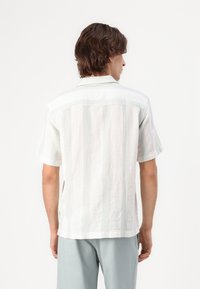 Camicia bianca a maniche corte con sottili strisce verticali verdi, tessuto testurizzato e colletto classico. I bordi sono rifiniti con cura.