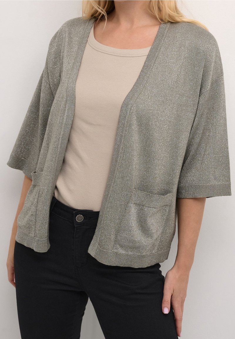 Femme portant un cardigan argenté métallique ouvert sur le devant avec poches avant, sur un haut beige côtelé sans manches et un pantalon noir.