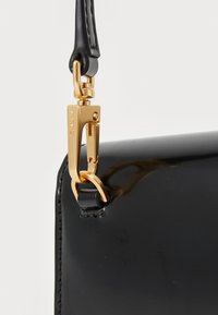 Sac en cuir verni noir avec un fermoir en métal doré et une finition lisse. La poignée est fixée par une sangle en cuir noir.
