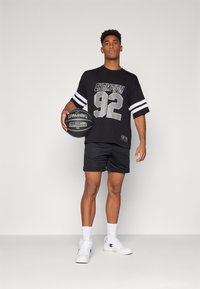 Camiseta negra de manga corta con la impresión "CHAMPION 92" y franjas blancas, combinada con pantalones cortos negros y zapatillas blancas, sosteniendo un balón de baloncesto negro.