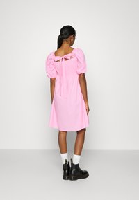 Vestido rosa de mangas bufantes com corpete ajustado e detalhe nas costas com laço, feito de material suave. Usado com botas de cano curto pretas e meias brancas.