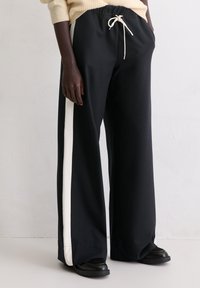 Osoba ubrana w czarne, szerokie spodnie z białymi paskami po bokach, kremowy sznurek i czarne buty, stojąca przed gładką ścianą.