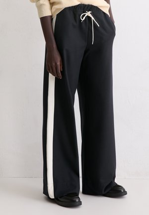 Person, der schwarze weit geschnittene Hosen mit weißen Seitenstreifen, eine cremefarbene Kordel und schwarze Schuhe trägt, steht vor einer schlichten Wand.