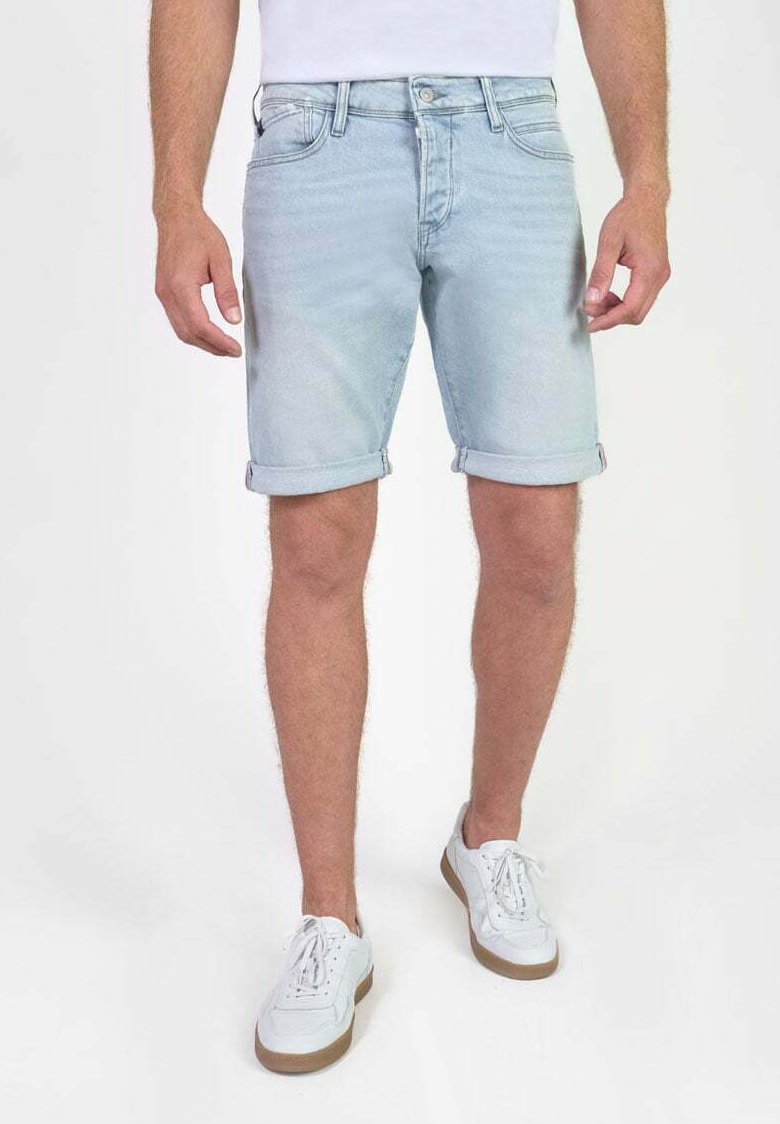 Man in lichtblauwe opgerolde denimshorts en witte sneakers, staand tegen een effen witte achtergrond.